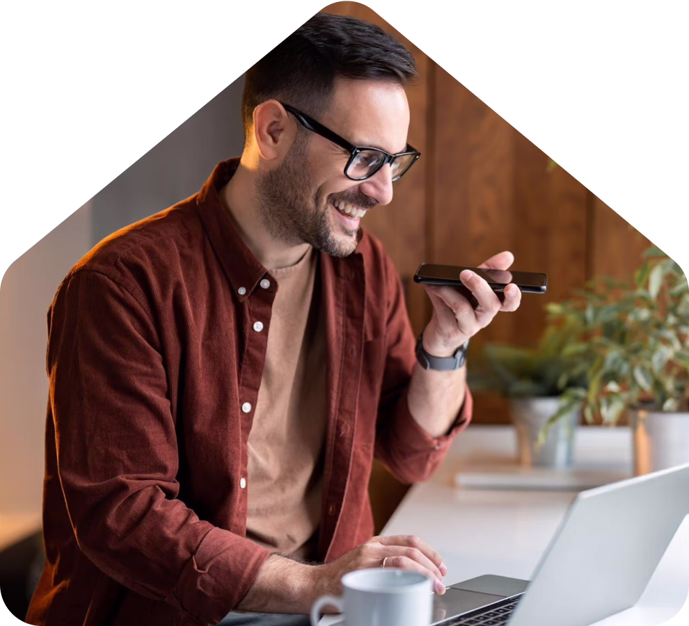 A man working on a laptop and holding a phone at a desk in a bright modern office with natural light and indoor plants
